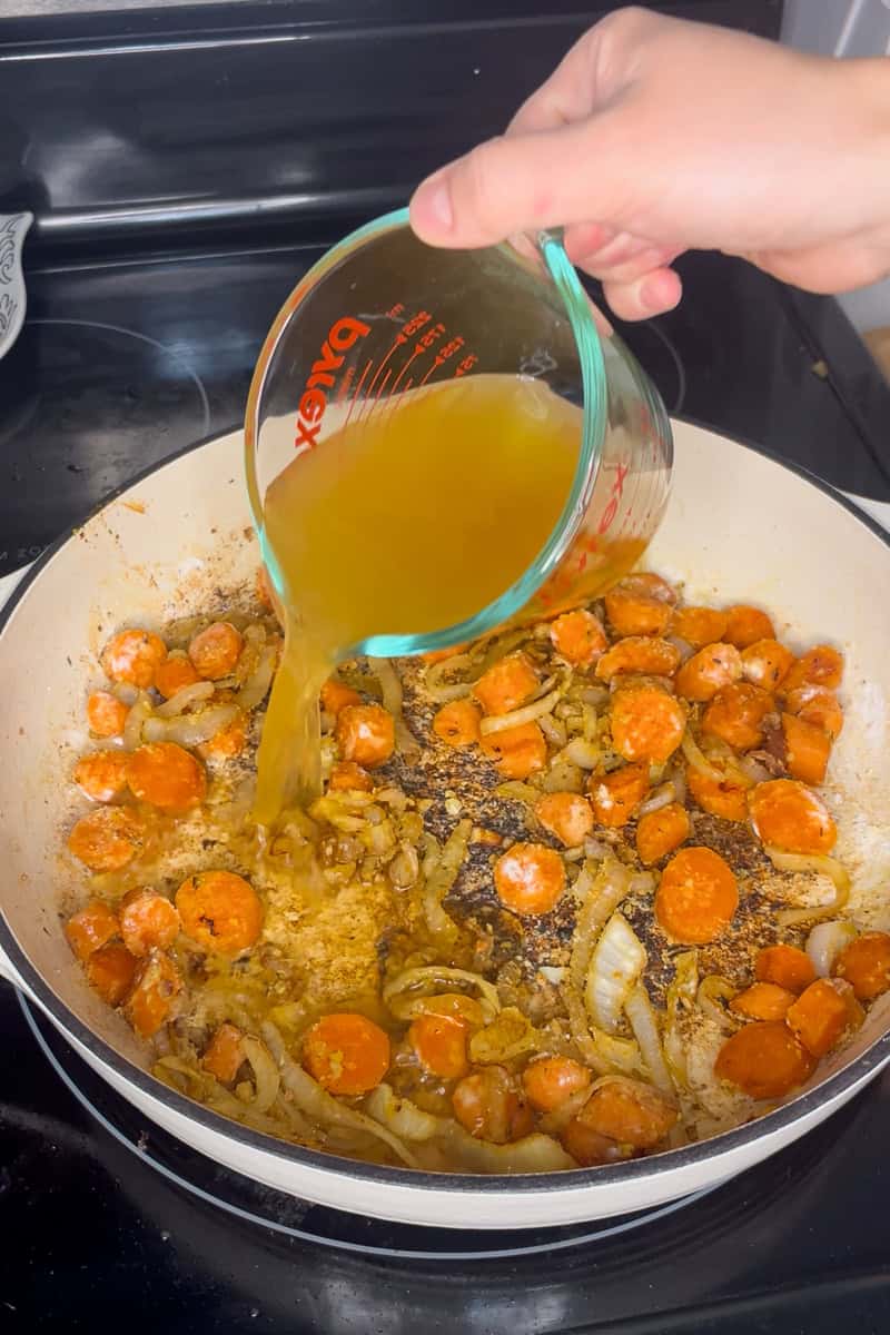 Pouring chicken broth into a pan with carrots and onions to deglaze for apple cider sauce.