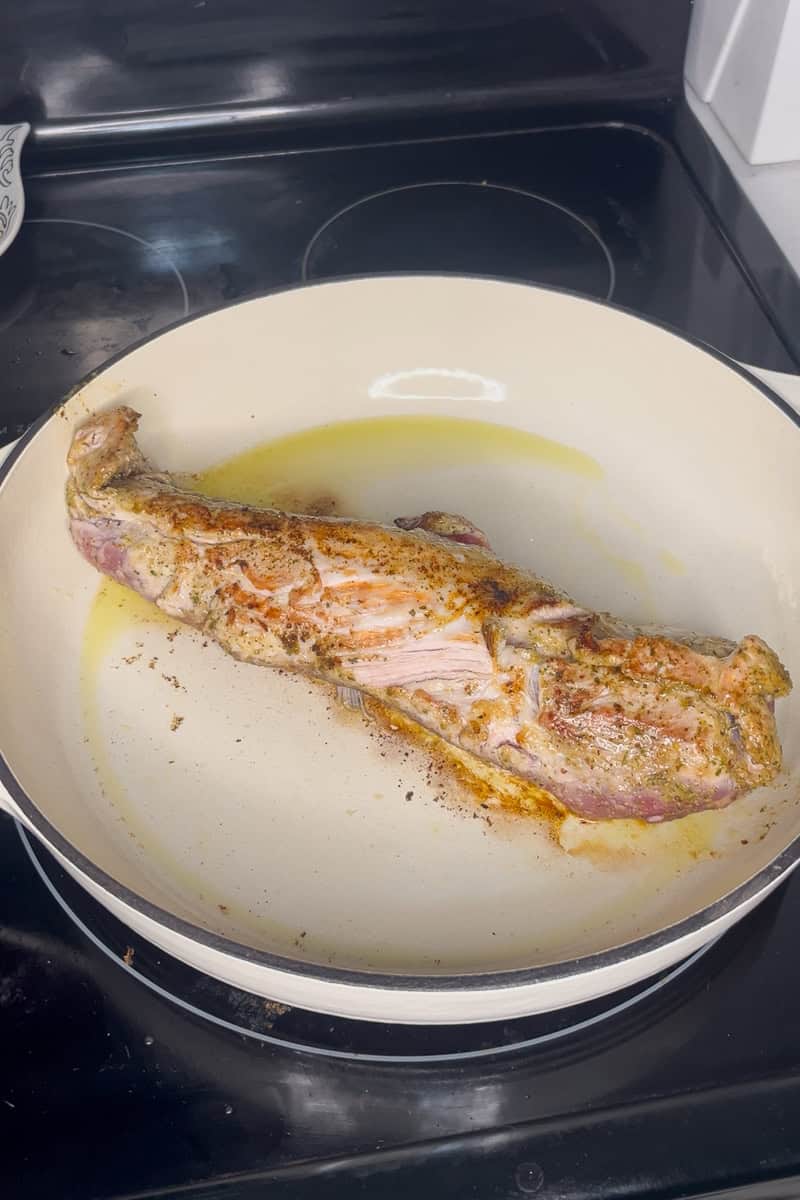 Pork tenderloin searing in a white braiser pan with olive oil on the stove.