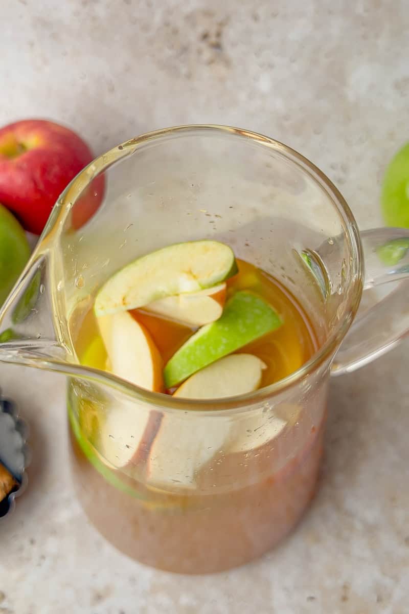 A glass pitcher filled with sliced red and green apples soaking in golden apple cider sangria.