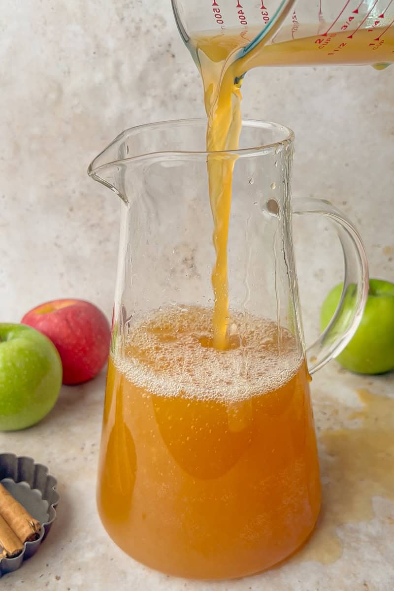 Apple cider being poured into a clear glass pitcher surrounded by fresh apples and cinnamon sticks.