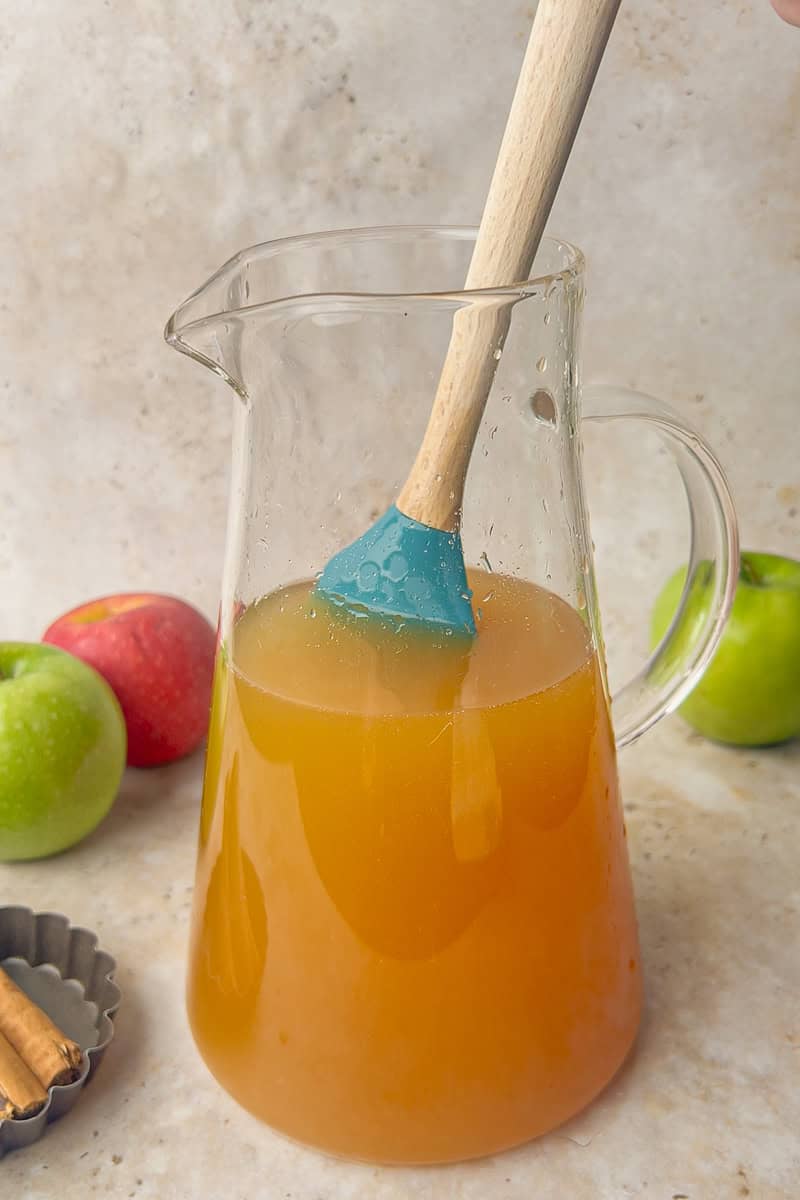 A wooden spoon stirring apple cider sangria in a glass pitcher with apples and cinnamon nearby.