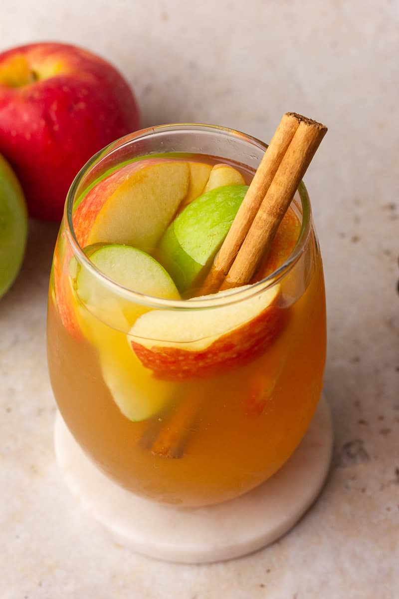 A glass of apple cider sangria filled with red and green apple slices and garnished with a cinnamon stick, sitting on a light surface.