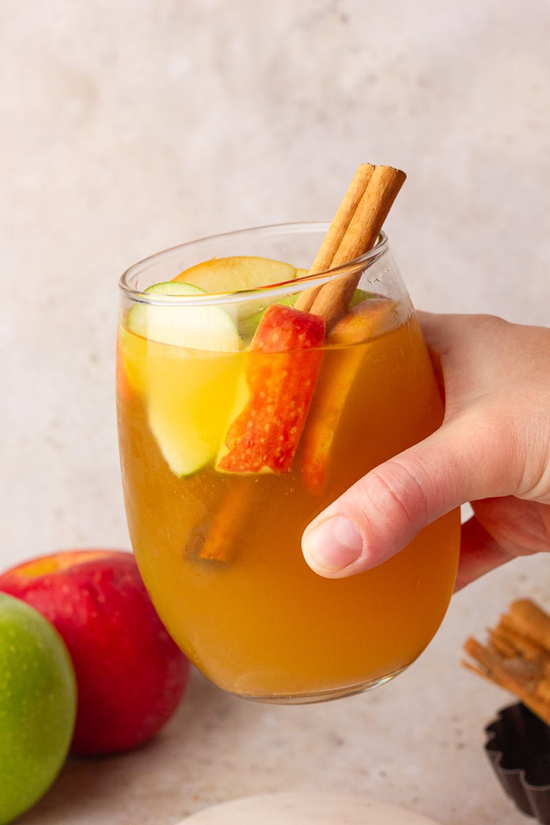 Hand holding a glass of apple cider sangria garnished with apple slices and a cinnamon stick.