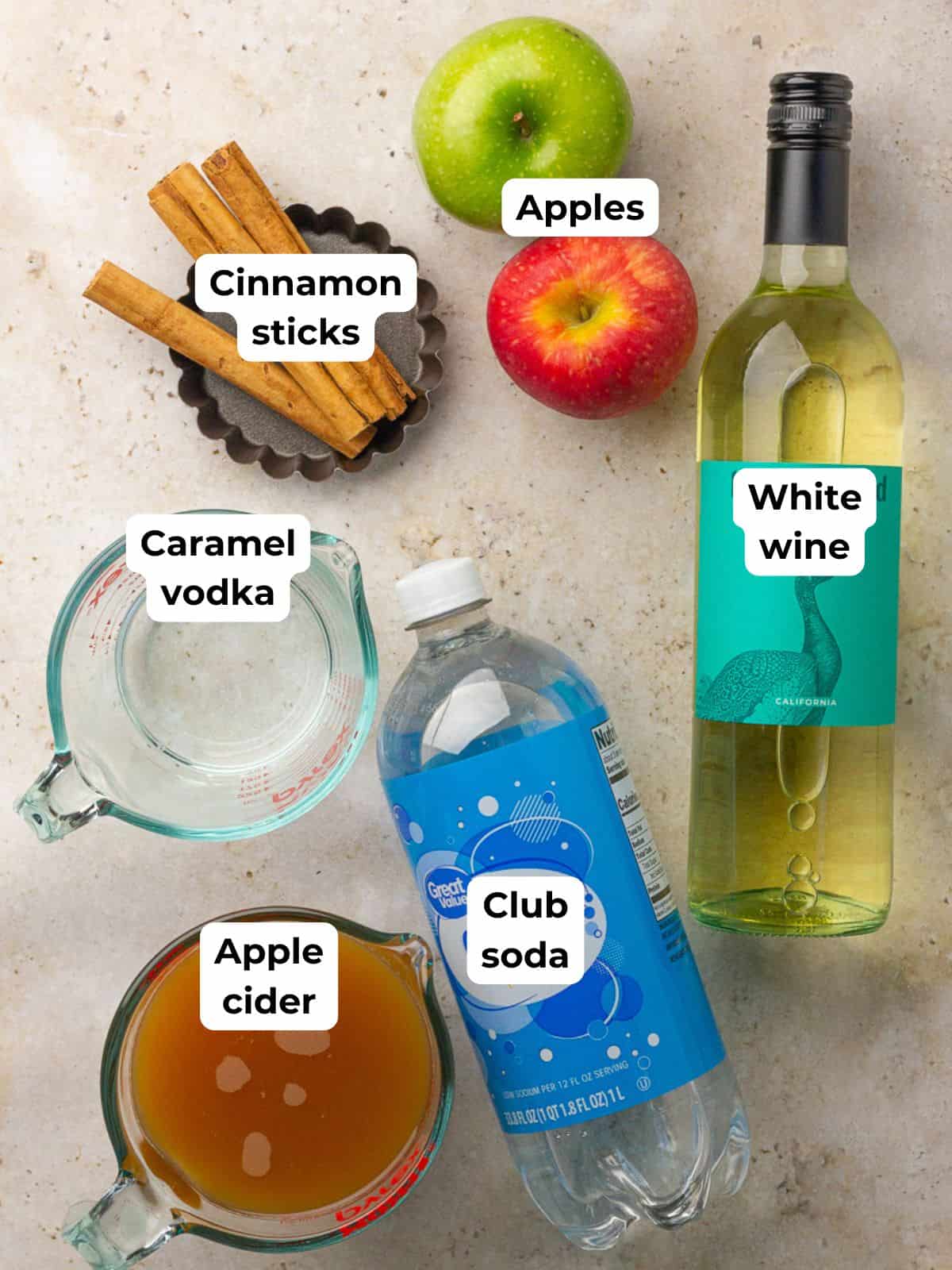 Overhead shot of ingredients for apple cider sangria including wine, apple cider, caramel vodka, apples, cinnamon sticks, and club soda.