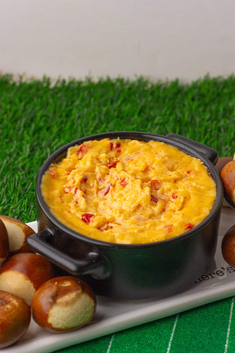 A small black bowl filled with hot baked pimento cheese dip surrounded by soft pretzel bites on a serving tray.