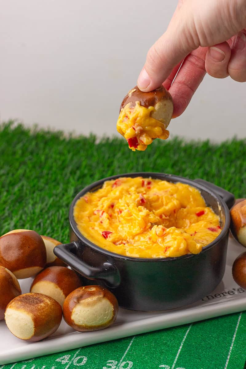 A hand dipping a pretzel bite into a bowl of creamy baked pimento cheese dip on a green football-themed surface.