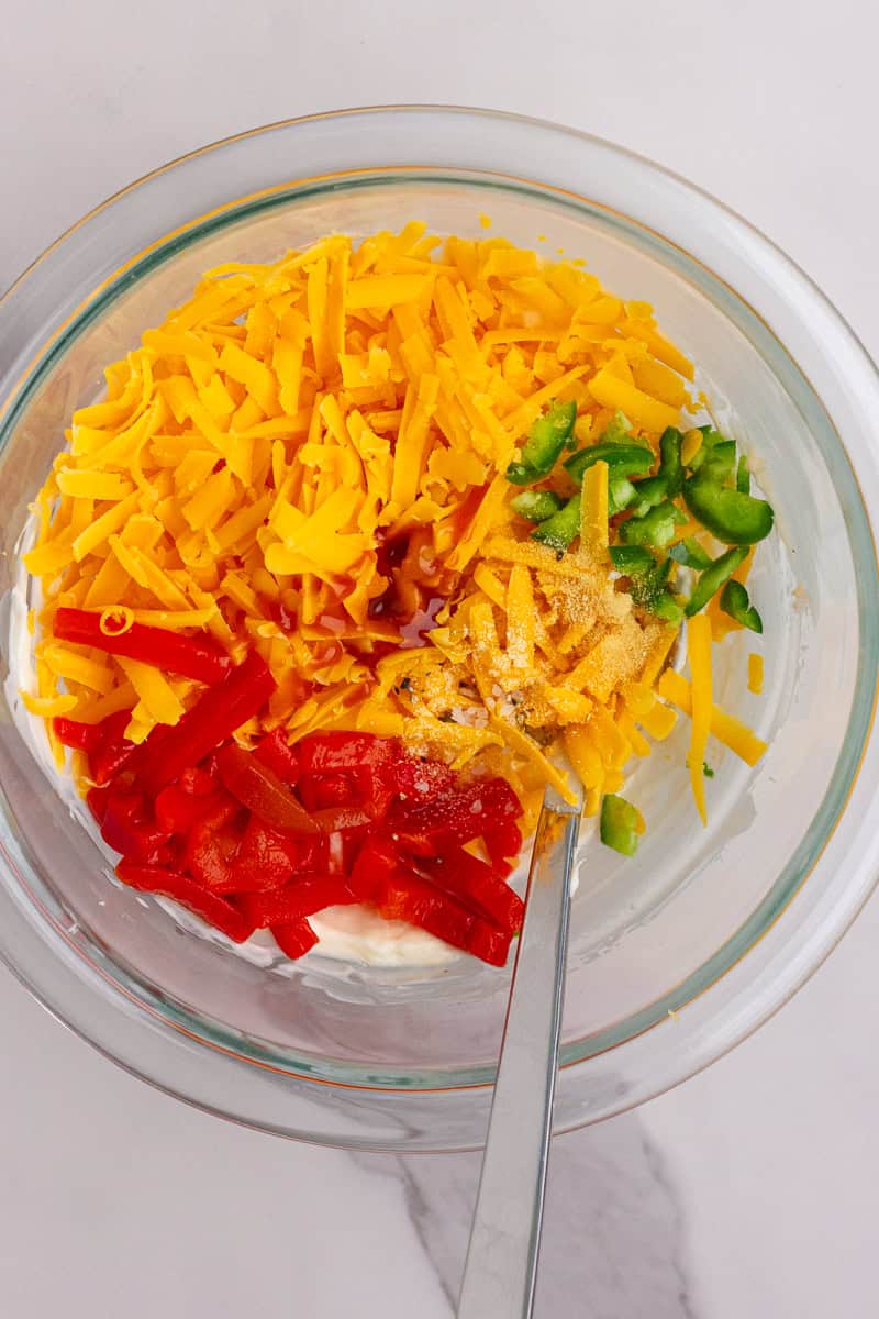 A glass bowl filled with shredded cheese, diced pimentos, jalapeño slices, and seasonings ready to be mixed for baked pimento cheese dip.