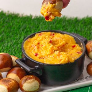 A hand dipping a soft pretzel bite into a bowl of hot, melty baked pimento cheese dip on a football-themed serving tray.