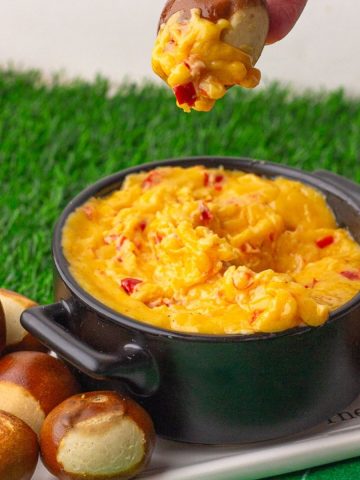 A hand dipping a soft pretzel bite into a bowl of hot, melty baked pimento cheese dip on a football-themed serving tray.