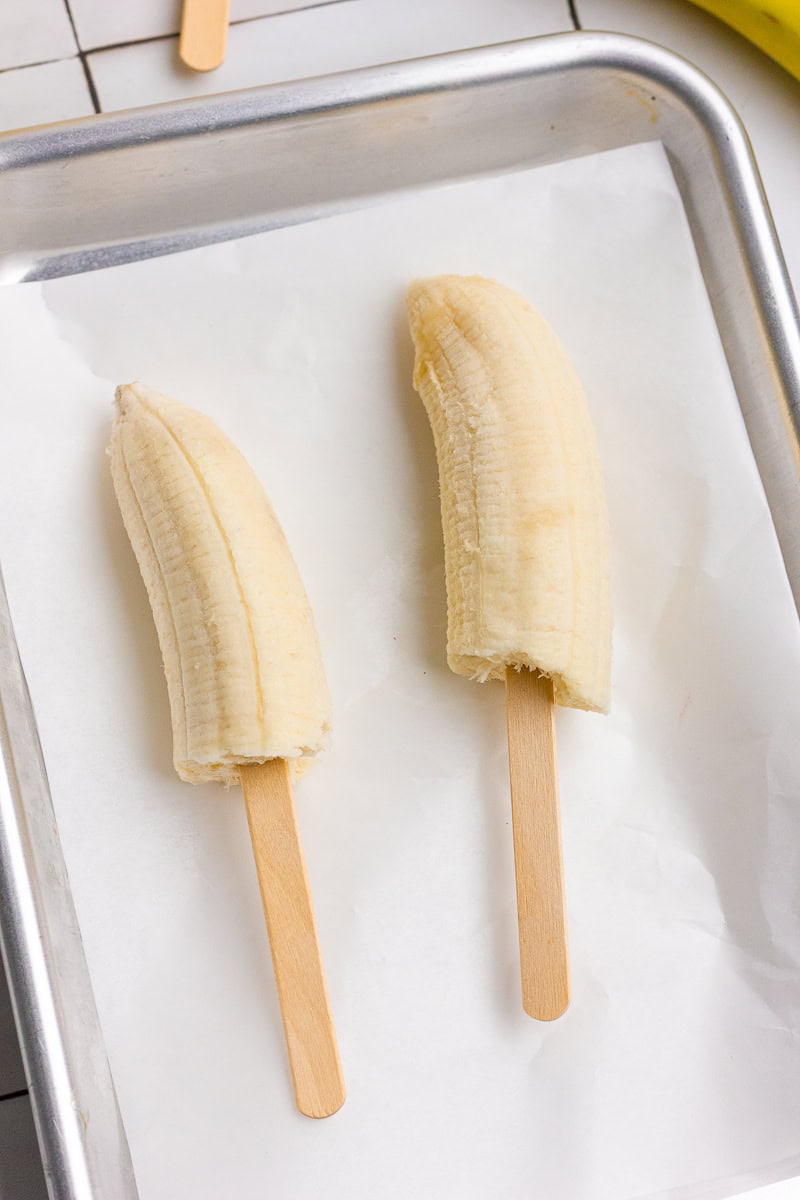 Two peeled banana halves with popsicle sticks on a parchment-lined baking sheet ready for dipping.