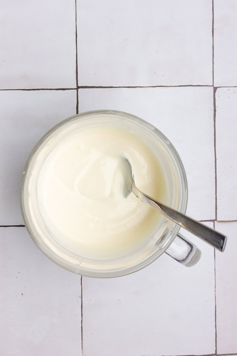 A clear bowl filled with melted white chocolate and a spoon resting inside on a tiled counter.