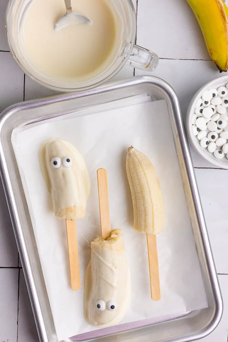 Partially dipped bananas with candy eyes being added on a tray next to melted white chocolate.