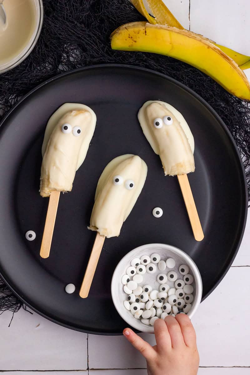 Close-up of a hand reaching for candy eyes next to three white chocolate-covered boonanas on a plate.