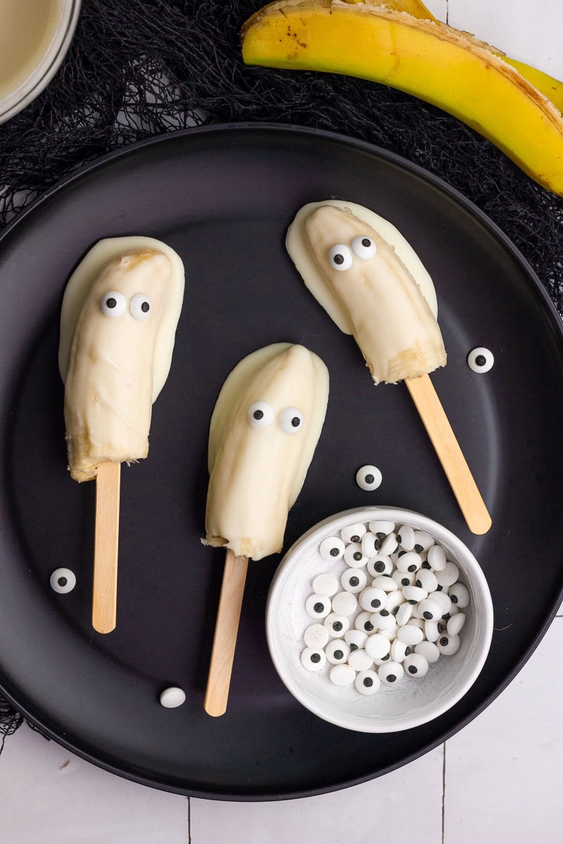 Three finished boonanas with candy eyes sitting on a black plate surrounded by extra candy eyes.
