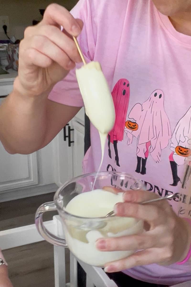A person wearing a pink ghost-themed Halloween shirt dipping a banana into melted white chocolate.