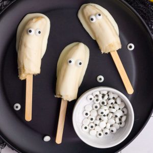 Finished white chocolate-covered boonanas with candy eyes on a black plate beside a bowl of candy eyes.