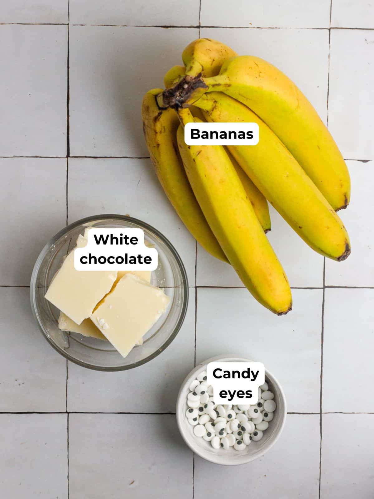 A bunch of bananas, a bowl of white chocolate, and candy eyes displayed on a white tile surface.