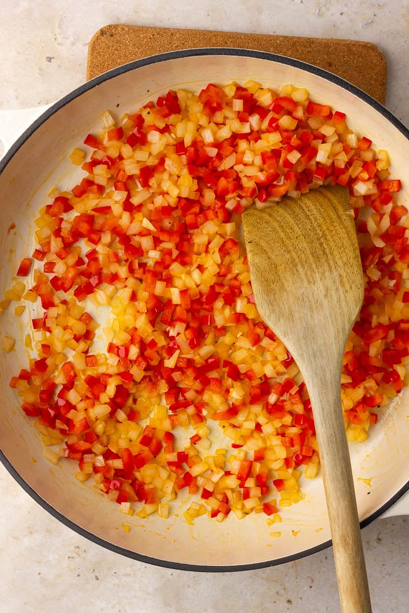 Diced onion and red bell pepper are sautéing in a large skillet with a wooden spatula.