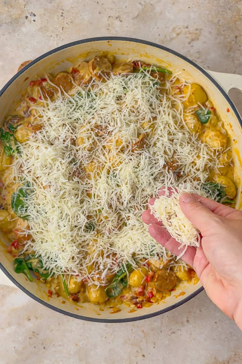 A hand sprinkles shredded mozzarella over the simmered gnocchi and spinach before the final bake.