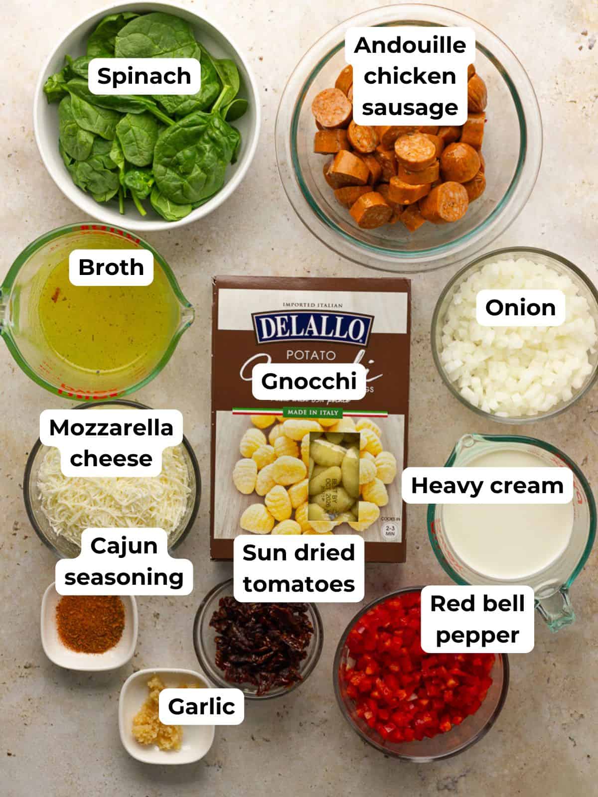 Measured ingredients for Cajun gnocchi—gnocchi, broth, andouille chicken sausage, onion, red bell pepper, spinach, garlic, sun-dried tomatoes, Cajun seasoning, heavy cream, and shredded mozzarella—are arranged around a box of gnocchi.