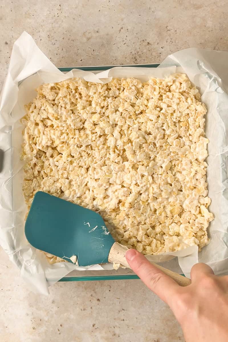 A blue spatula presses the Rice Krispie mixture into a parchment-lined pan.
