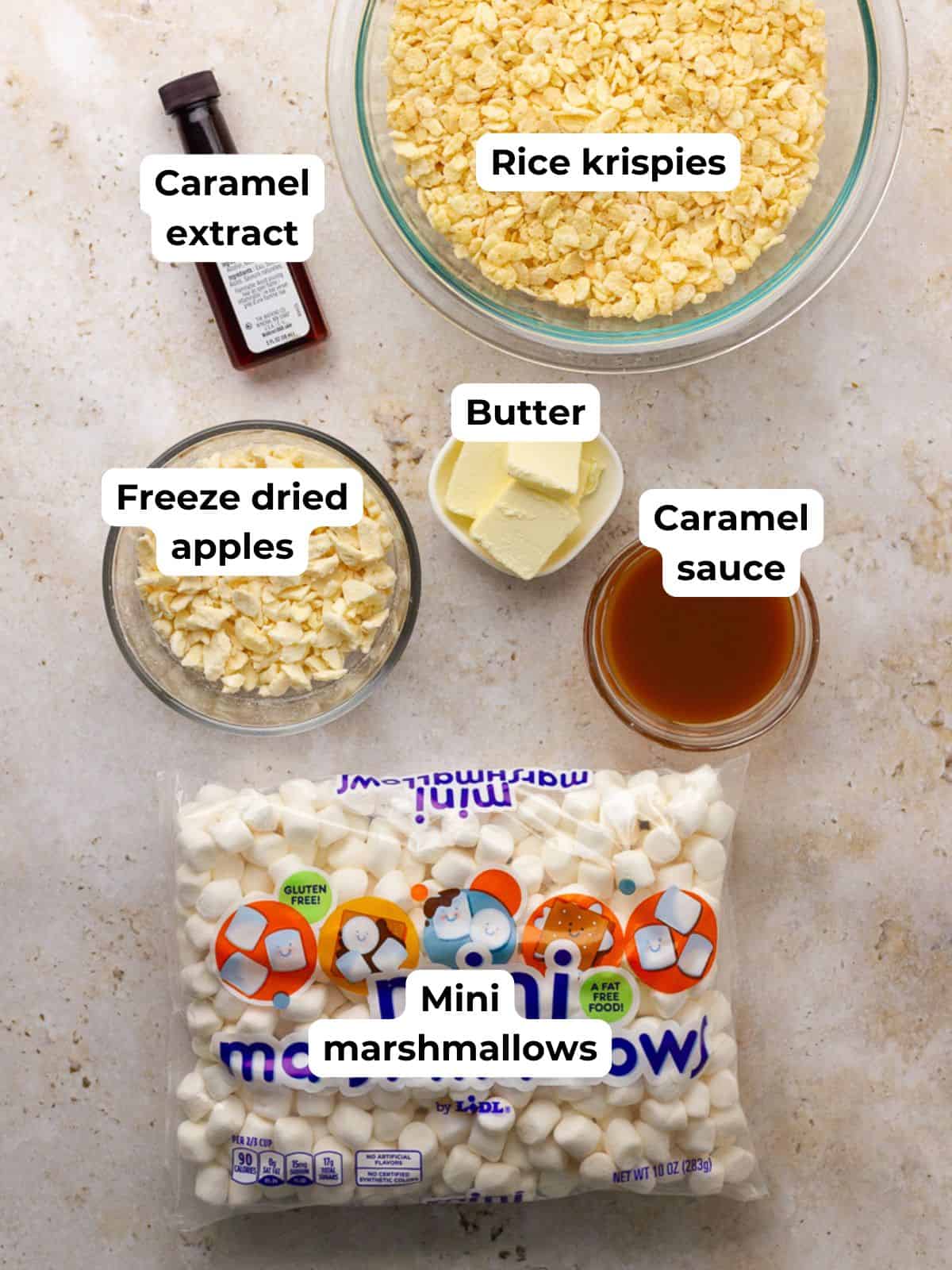 A flat lay shows bowls of rice cereal, mini marshmallows, butter, caramel extract, caramel sauce, and freeze-dried apples.