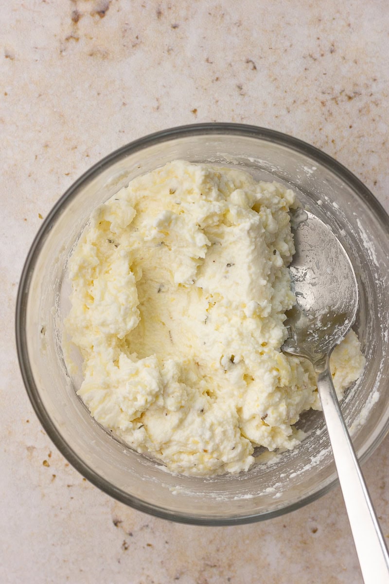 A glass bowl shows a creamy ricotta cheese mixture with Italian seasoning and a spoon.