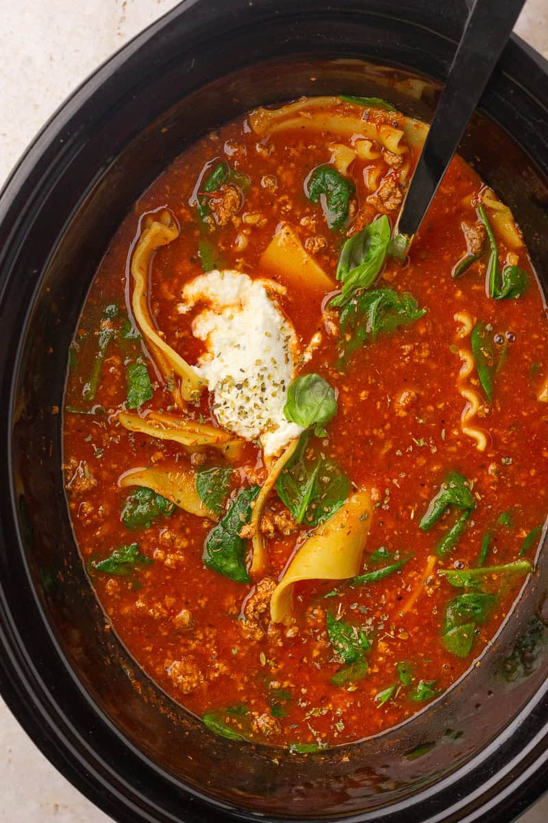 A slow cooker holds red lasagna soup with spinach, noodle pieces, and a dollop of ricotta on top.