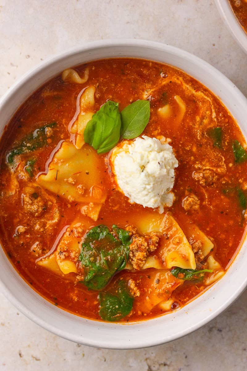 A close-up bowl of lasagna soup shows noodle pieces, spinach, crumbled meat, a ricotta scoop, and fresh basil.