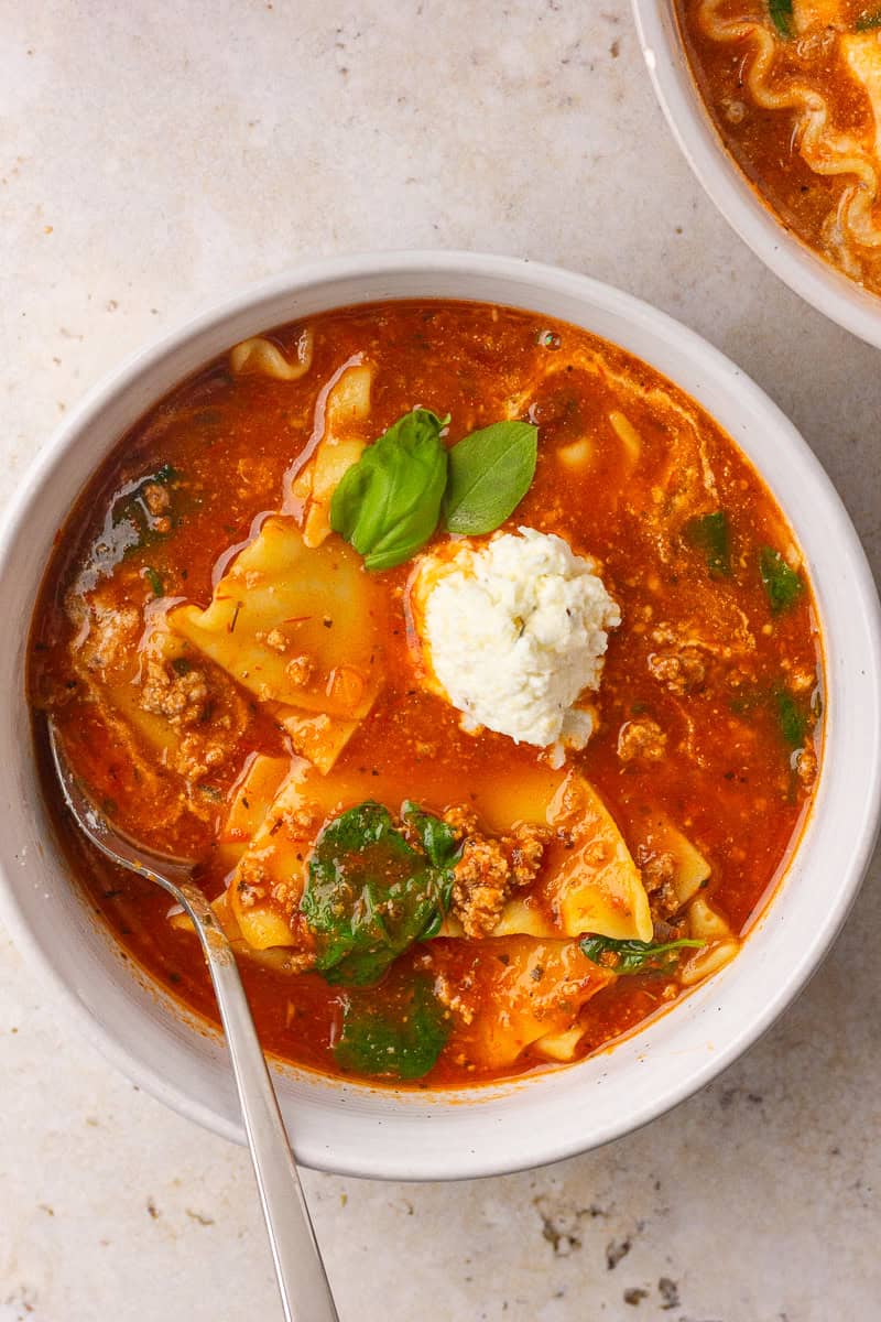 A white bowl of lasagna soup shows noodles, spinach, a ricotta scoop, fresh basil, and a spoon.