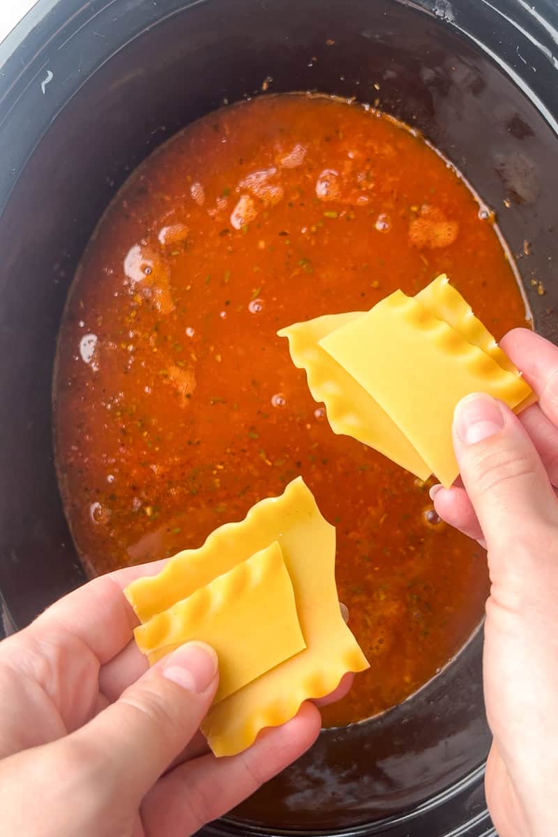 Hands hold broken lasagna noodles over a slow cooker filled with the red soup base.