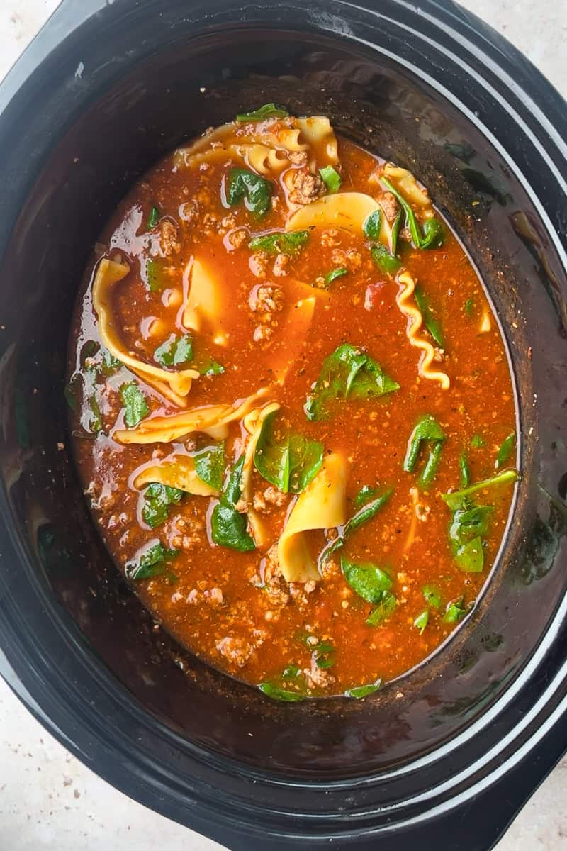 A slow cooker shows lasagna soup simmering with noodles, spinach, and crumbled meat.