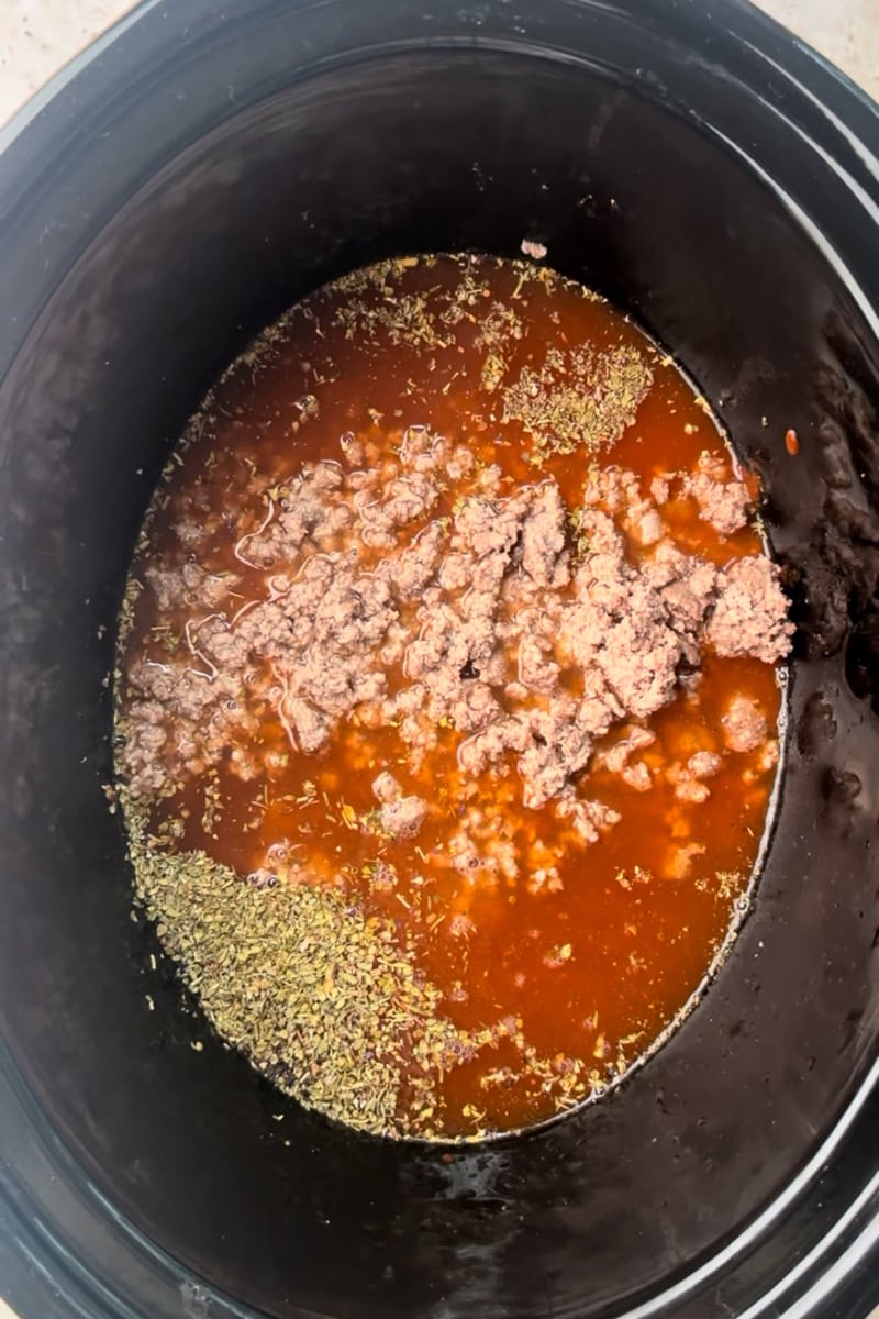 The slow cooker contains marinara, browned meat, and Italian seasoning before being stirred together.
