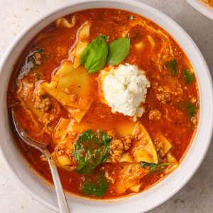 A bowl of lasagna soup features noodles, spinach, a ricotta dollop, fresh basil, and a spoon.