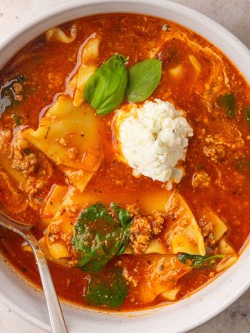 A bowl of lasagna soup features noodles, spinach, a ricotta dollop, fresh basil, and a spoon.