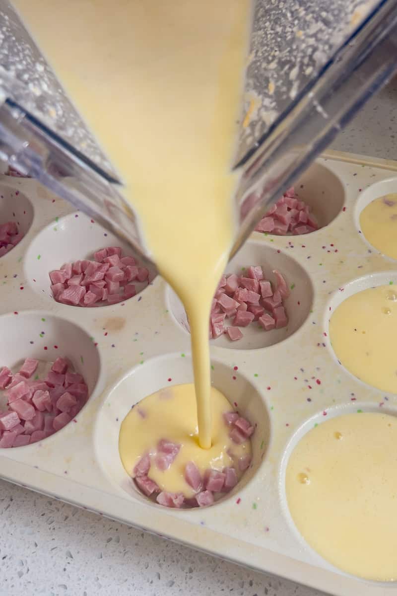 Egg mixture being poured from a blender into muffin tins filled with diced ham.