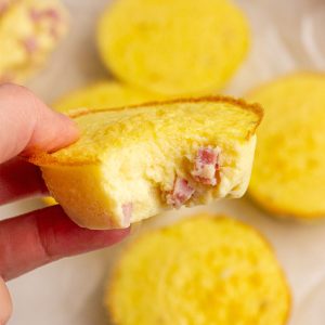 A hand holding a fluffy ham and cheese egg bite with more bites on parchment in the background.