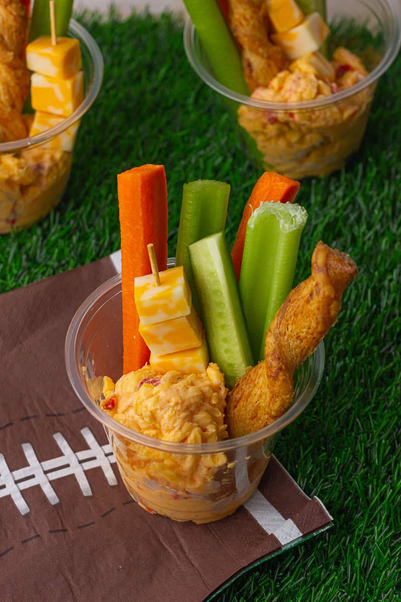 Single-serve jalapeno pimento cheese cups filled with dip, veggies, cheese cubes, and pretzel twists on a football napkin.