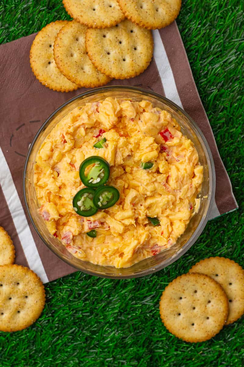 Bowl of jalapeno pimento cheese topped with sliced jalapeños and surrounded by round crackers on a football napkin.