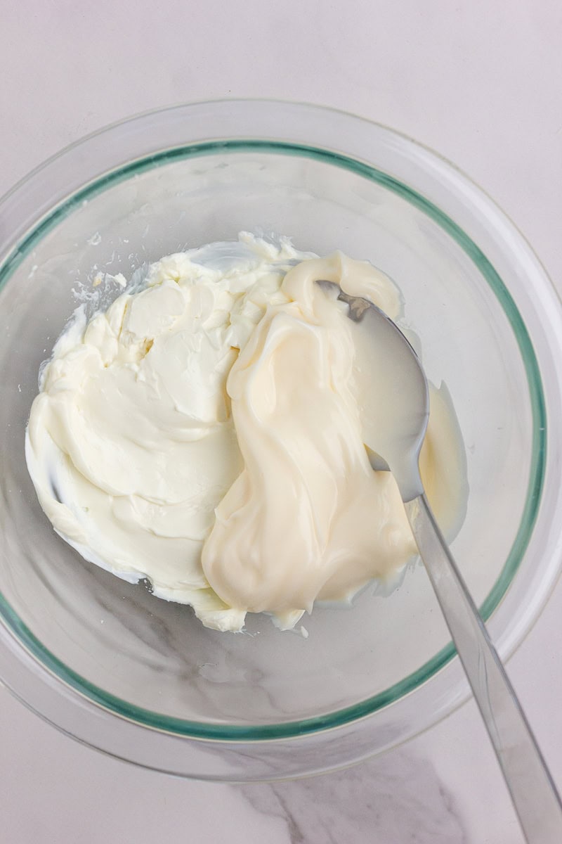 Mixing bowl with softened cream cheese and mayonnaise being combined with a spoon.