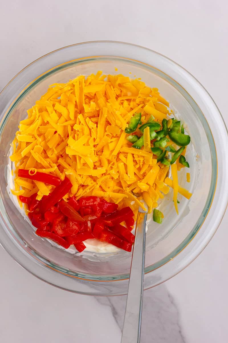 Mixing bowl filled with shredded cheddar, diced jalapeños, and chopped pimentos before stirring together.