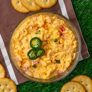 Overhead view of creamy jalapeno pimento cheese topped with sliced jalapeños and served with crackers on turf.