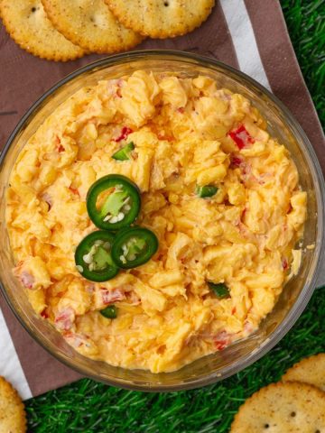 Overhead view of creamy jalapeno pimento cheese topped with sliced jalapeños and served with crackers on turf.