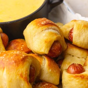 Close-up of baked mini pretzel dogs with golden tops and coarse salt next to a bowl of cheese dip.