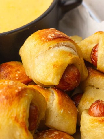 Close-up of baked mini pretzel dogs with golden tops and coarse salt next to a bowl of cheese dip.