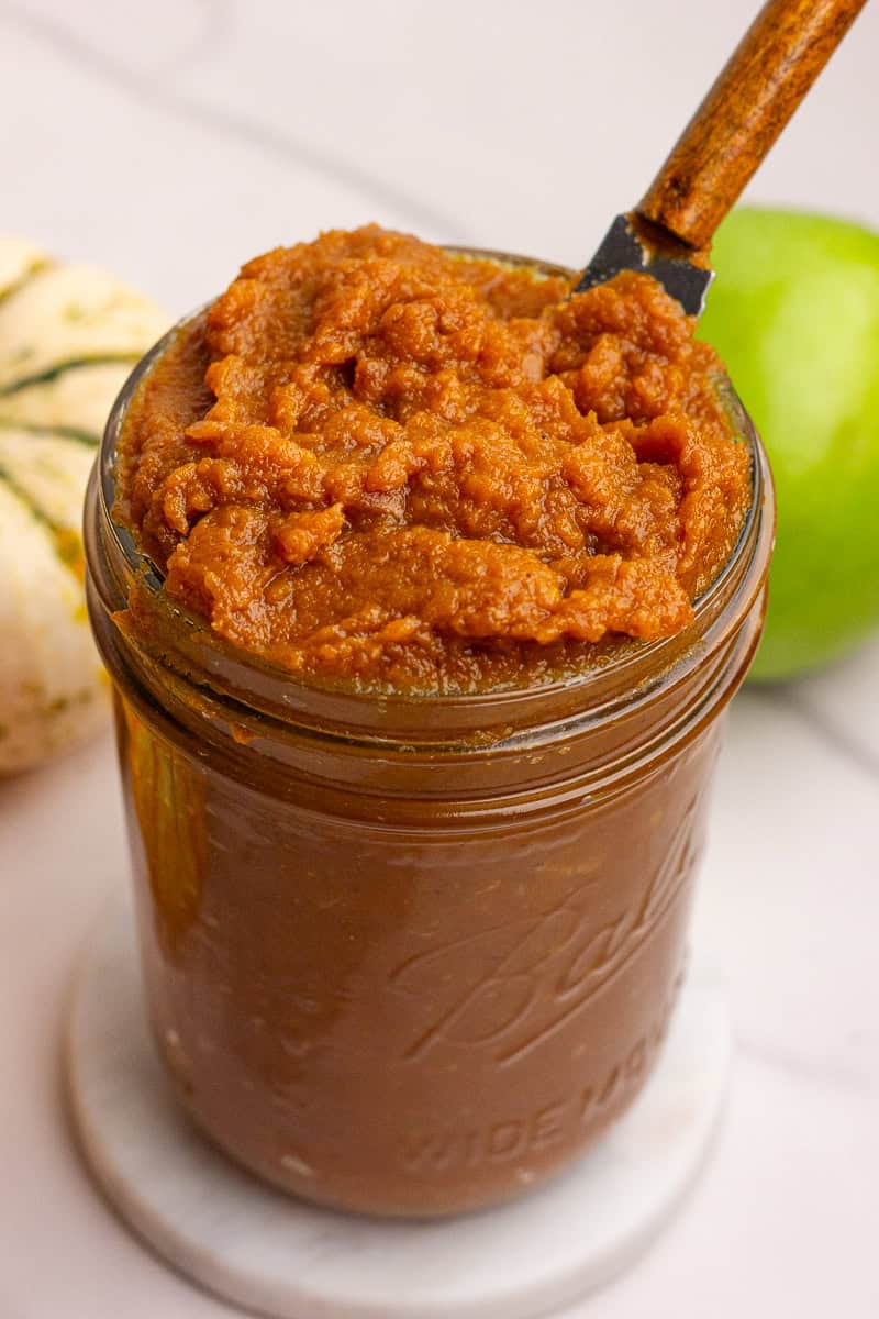 A jar of finished pumpkin apple butter with a knife scooping from the top, surrounded by fall produce.