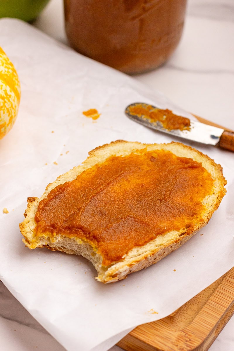 Slice of bread spread with pumpkin apple butter on parchment paper next to a butter knife.