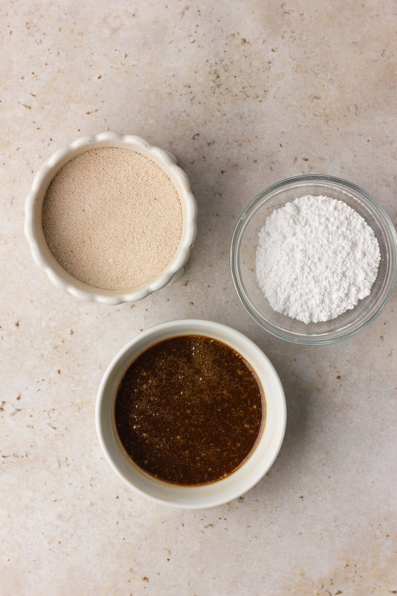 Three small bowls with cinnamon sugar, powdered sugar, and maple glaze on a light countertop.