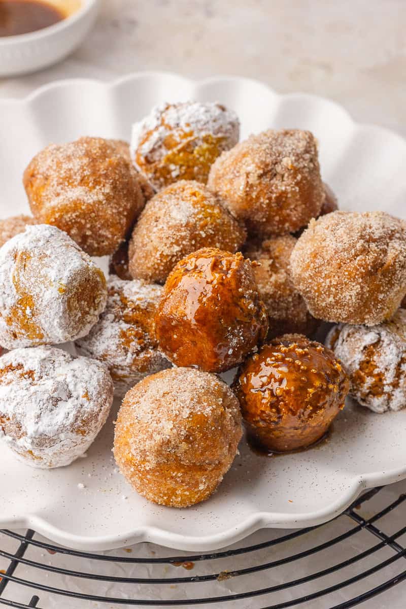 A pile of pumpkin donut holes on a white scalloped plate coated in cinnamon sugar, powdered sugar, and maple glaze.