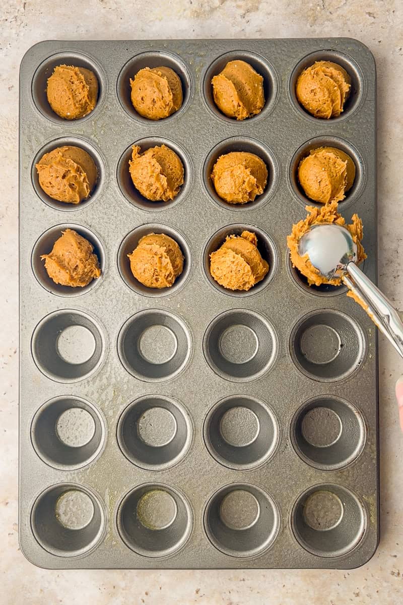 Overhead of a mini muffin pan partially filled with scoops of pumpkin batter as a small cookie scoop adds the next portion.